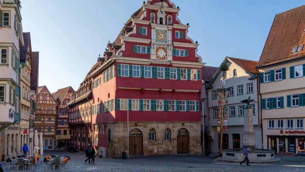 مشهد لمدينة ألمانية قديمة بمبنى تاريخي مميز بواجهة حمراء وساعة في الأعلى، مع شوارع ضيقة ومارة في الساحة.