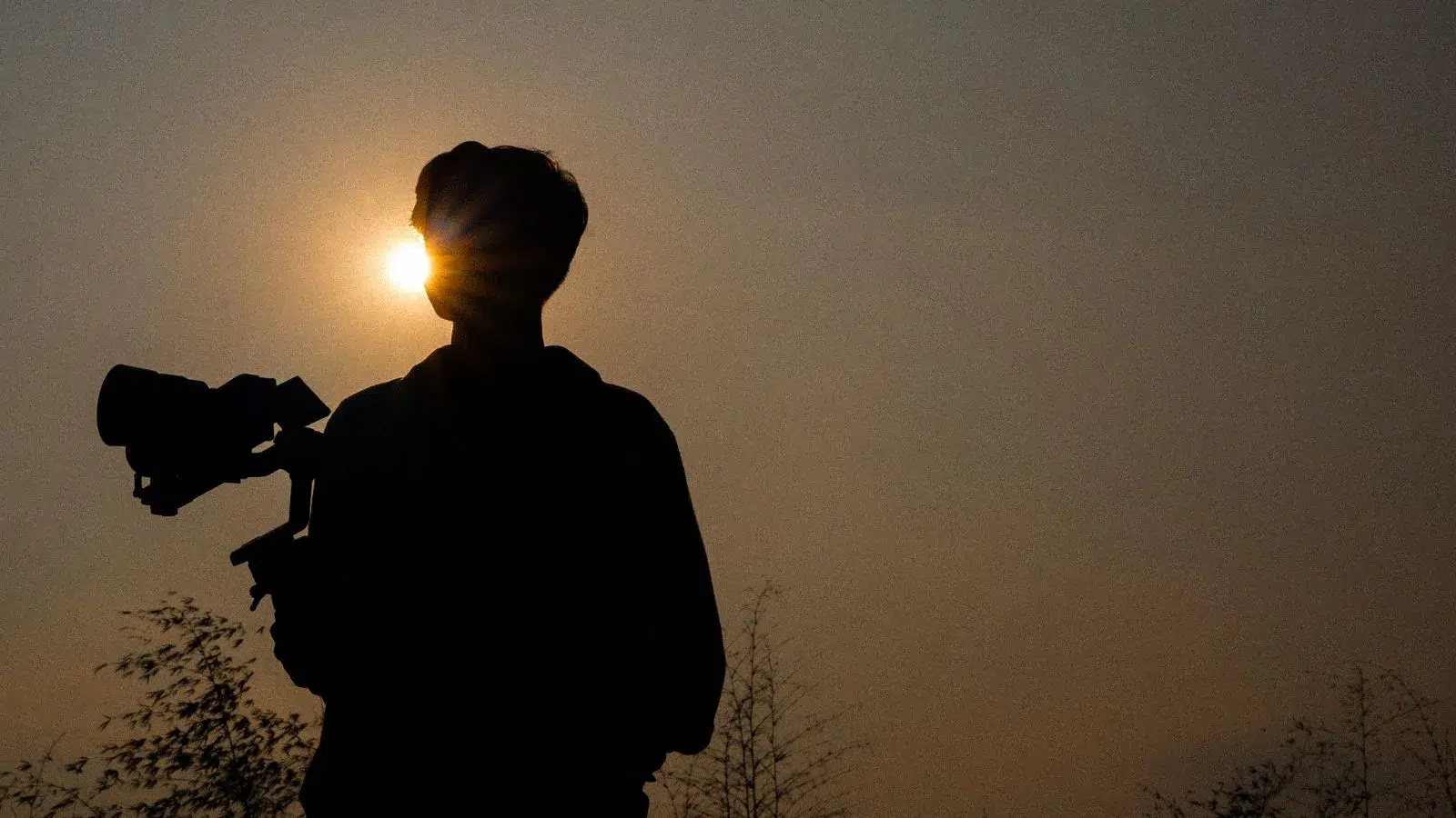 شخص يحمل كاميرا تصوير احترافية في وضعية ظل أمام ضوء الشمس، يرمز إلى فن صناعة الأفلام والتصوير السينمائي.