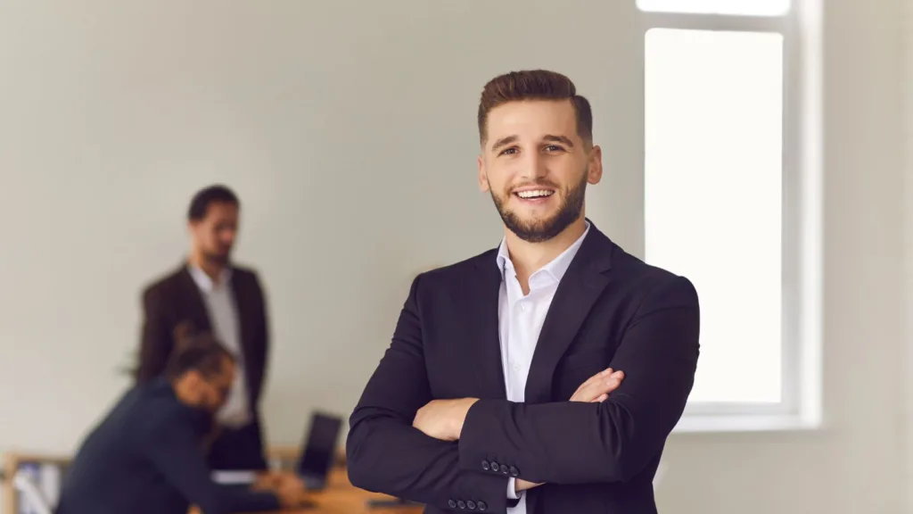 رجل يرتدي بدلة رسمية يقف مبتسمًا في مكتب مع زملاء يعملون في الخلفية.